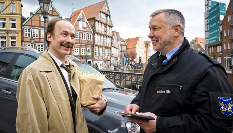Chlodwig Pullmann (Ulrich Noethen) versucht mit einem Lächeln dem Strafzettel des Polizisten (Wilfried Mahler) zu entkommen. – Bild: NDR/​Svenja von Schulzendorff