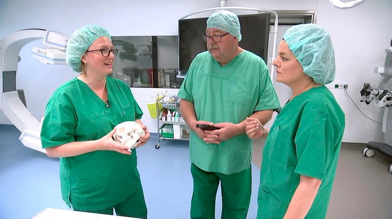 Dr. Heike Israel-Willner (li.) von der Kopfschmerzambulanz der Charité Berlin erklärt einem Patienten (Mitte) mit Cluster-Kopfschmerzen im Operationssaal der Neurochirurgie ein neues Verfahren gegen die qualvollen Schmerzen. – Bild: SWR/​rbb/​Cornelia Fischer-Börold