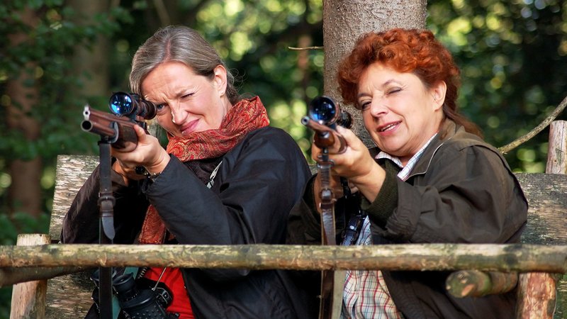 Angela Klopp (Sybille Schedwill, links) und Gundi Hugl (Monika Baumgartner) versuchen, einen Hirsch zu erlegen. Doch worauf genau sie da anlegen, wissen sie noch nicht. – Bild: BR/​Simon Weber