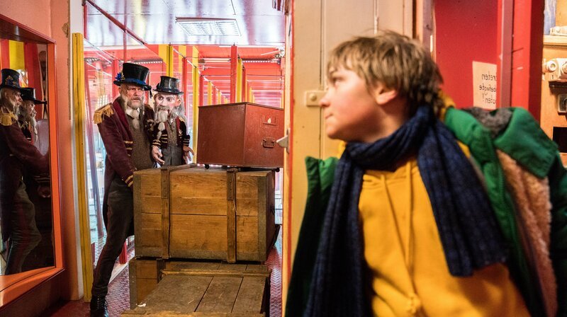 Frido (Luis Vorbach) versteckt sich hinter dem Schrank, während der Spiegelkabinettbesitzer (Butz Buse) eine Kiste verschiebt. – Bild: MDR/​VIAFILM/​Marc Reimann