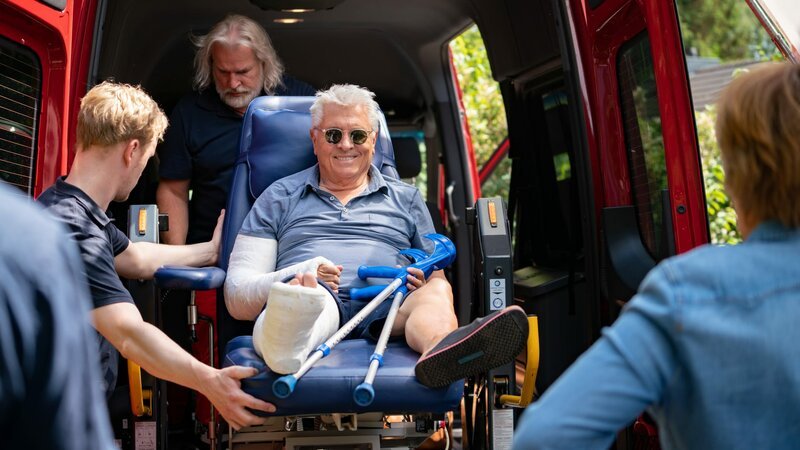 Volker (Henry Hübchen, M.) wird vom Krankenwagen „geliefert“. Er hat einen Arm und einen Bein in Gips. Sanitäter (Jasper Diedrichsen, l., Frank Smolarek, 2.v.l.) heben ihn aus dem Wagen. – Bild: ZDF und Georges Pauly.