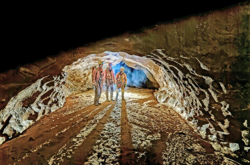 Drei Höhlenforscher stehen in der Riesending-Höhle, der größten bekannten Schachthöhle Deutschlands, unter einem gigantischen Steinbogen. – Bild: Filmwelt Verleihagentur/​Dr. Wolfgang Zillig