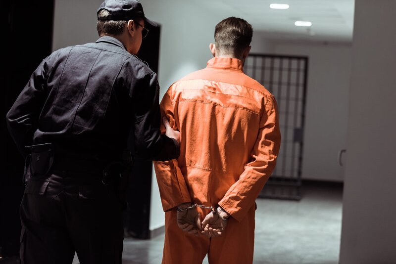 Rückansicht eines Gefängnisbeamten, der einen Gefangenen in Handschellen durch den Korridor führt – Bild: Shutterstock /​ Shutterstock /​ Copyright (c) 2018 LightField Studios/​Shutterstock. No use without permission.
