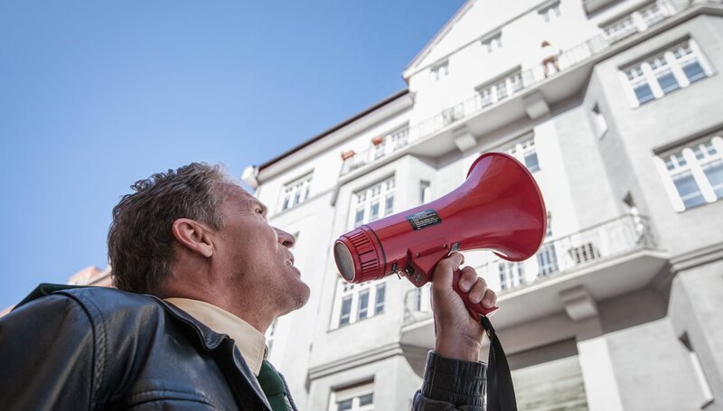 Franz Hubert (Christian Tramitz) spricht über das Megaphon mit der lebensmüden Anna Schmitt, die von ihrem Balkon springen will. – Bild: ARD/​Chris Hirschhäuser