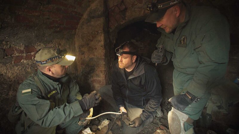 L-R: Josh Gates und die Ermittler Pawel Piatkiewicz und Robert Kubiszekski entdecken in dem Nazitunnel einen verfallenen Bilderrahmen aus Holz und Glas. – Bild: Warner Bros. Discovery