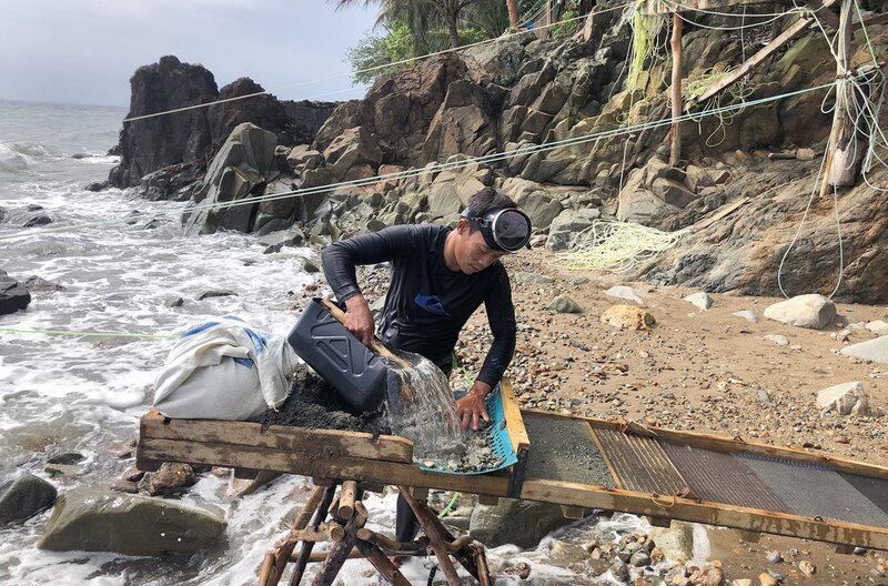 Ein Goldtaucher am Strand bei Pinut-An siebt Sand aus Säcken: Das darin enthaltene Gold bleibt an der Matte hängen. – Bild: Carsten Stormer /​ © Carsten Stormer