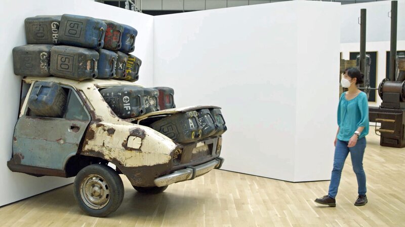 Eine Ausstellungsbesucherin vor dem Kunstwerk Elf rien à foutre (Künstler: Romuald Hazoumè): ein halbes verrostetes Auto vollgefüllt mit Benzinkanistern, die auch auf dem Dach befestigt sind; das Auto verschwindet zur Hälfte in der Wand. – Bild: ORF/2022 NAVIGATOR FILM & Vive LaDok Eine Ausstellungsbesucherin vor dem Kunstwerk Elf rien à foutre (Künstler: Romuald Hazoumè): ein halbes verrostetes Auto vollgefüllt mit Benzinkanistern, die auch auf dem Dach befestigt sind; das Auto verschwindet zur Hälfte in der Wand. – Bild: ORF/2022 NAVIGATOR FILM & Vive LaDok