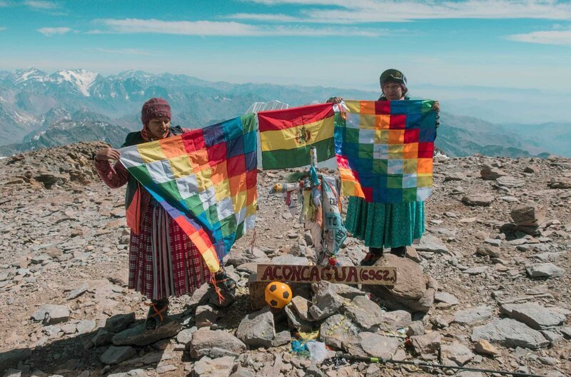 Die Cholitas erklimmen den Berg in traditionellen Kleidern und mit ihrer Flagge – der Wiphala – als farbenfroher Ausdruck des Stolzes für das jahrhundertealte Erbe des Aymara-Volkes. – Bild: Arena Comunicacion /​ Die Cholitas erklimmen den Berg in traditionellen Kleidern und mit ihrer Flagge – der Wiphala – als farbenfroher Ausdruck des Stolzes für das jahrhundertealte Erbe des Aymara-Volkes.