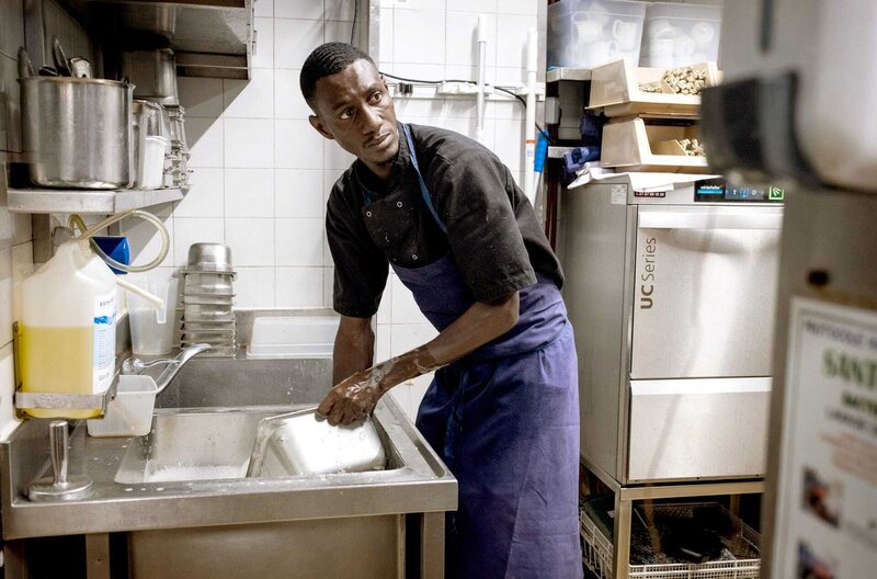 Immer zu Diensten – Bild: Camille Millerand /​ Makan Baradji arbeitet als Tellerwäscher in der Spülküche einer vornehmen Brasserie in der Nähe der Champs-Elysées. Als er die Stelle bekam, hatte er noch keine gültigen Ausweispapiere.