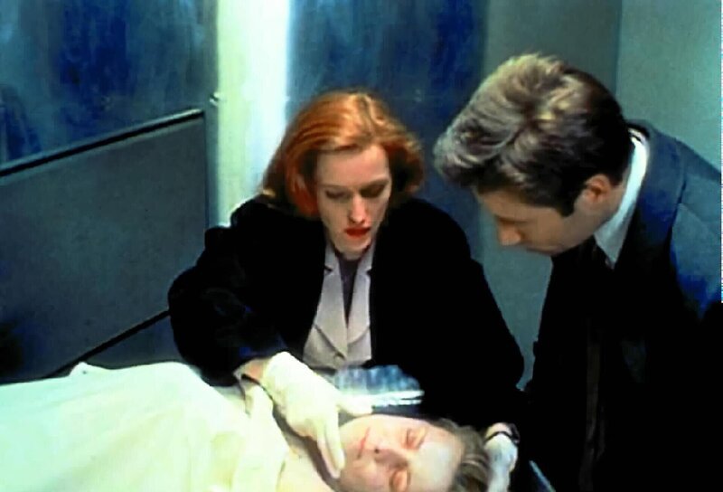 Scully (Gillian Anderson, l.) will Mulder (David Duchovny, r.) eine Phosphoressenz um Mund und Nase einer ermordeten Prostituierten zeigen, die sie bei ihrer ersten Untersuchung entdeckt hat. – Bild: SYFY