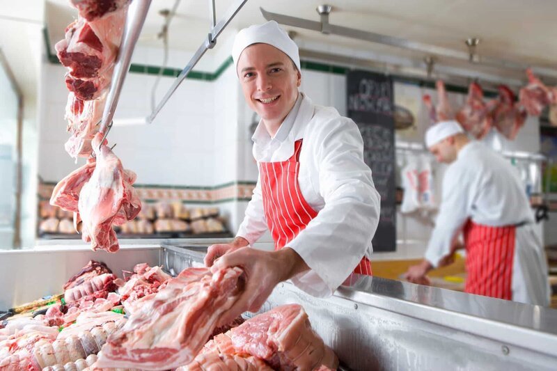 A young butcher smiling and holding meat near the camera in a meat shop. – Bild: Shutterstock /​ Shutterstock /​ Copyright (c) 2020 Juice Verve/​Shutterstock. No use without permission.