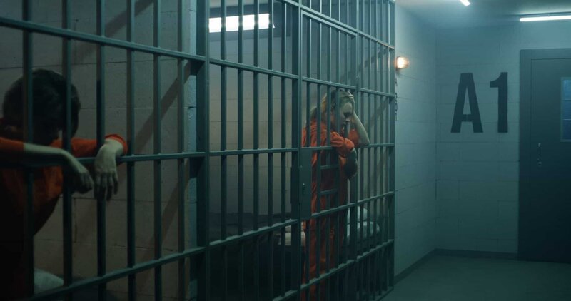 Eine weibliche Gefangene in orangefarbener Uniform steht hinter Metallgittern, eine andere sitzt auf dem Bett einer Gefängniszelle. – Bild: Copyright (c) 2023 Frame Stock Footage/​Shutterstock. No use without permission.