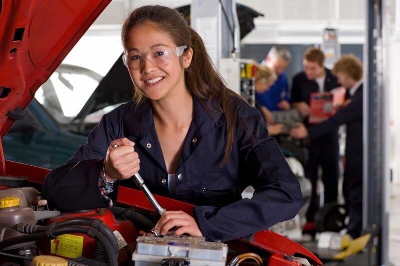 A horizontal view of a young girl in mechanic uniform and safety glasses repairing a car in the automotive training school – Bild: Shutterstock /​ Shutterstock /​ Copyright (c) 2020 Juice Verve/​Shutterstock. No use without permission.
