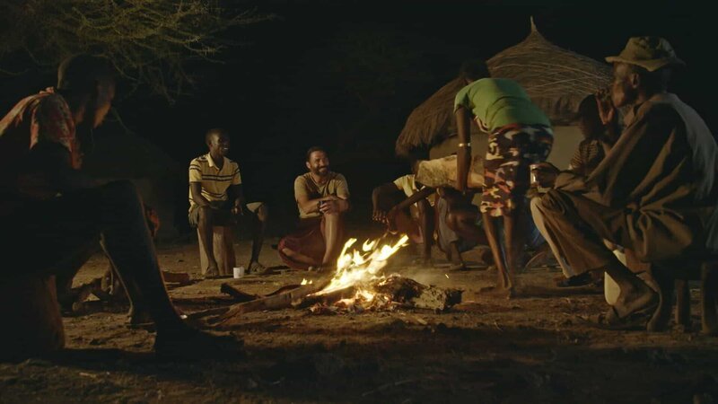 Hazen sitzt mit Einheimischen im Dorf am Lagerfeuer. (National Geographic für Disney) – Bild: National Geographic for Disney /​ Disney