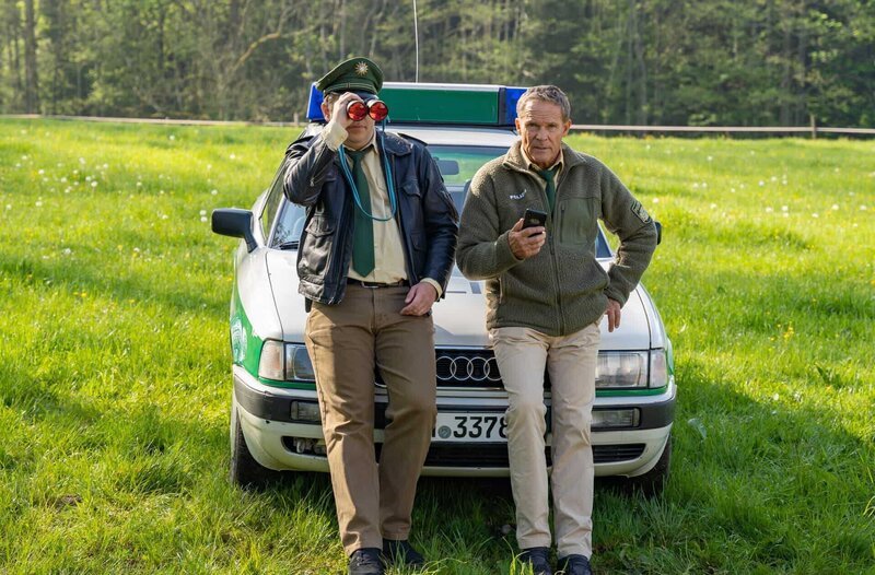 Hubert (Christian Tramitz, r.) und Riedl (Paul Sedlmeir, l.) lehnen am Streifenwagen und beobachten Girwidz. – Bild: ARD/​Thomas Neumeier