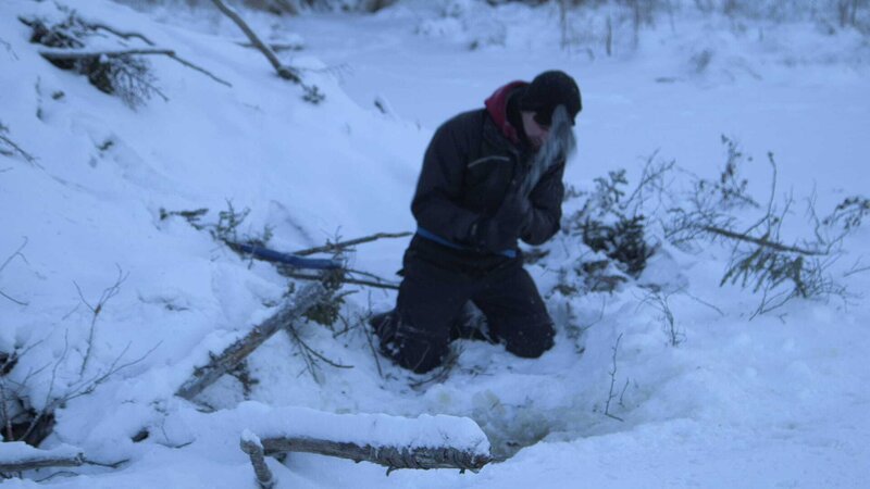 Kyle kneels in the snow surrounded by branches and sticks. – Bild: Discovery Communications