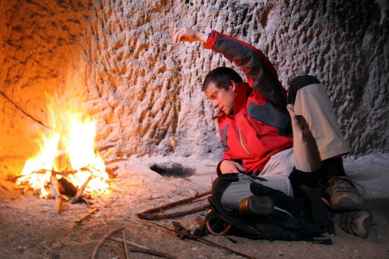 Bear Grylls lays in front of burning fire in man-made cave. – Bild: Discovery Communications