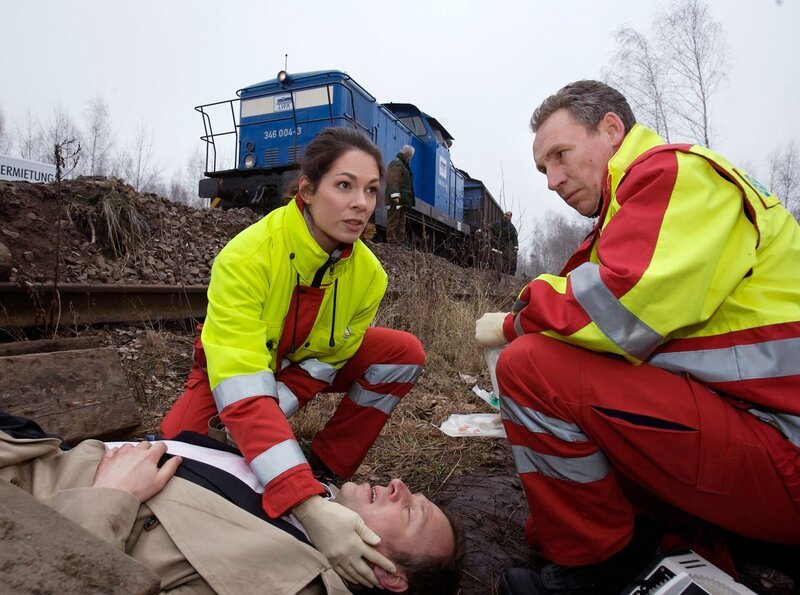Dr. Elena Eichhorn (Cheryl Shepard, links) trifft mit einem Kollegen (Komparse, rechts) am Unfallort ein. Dr. Ralph Groher (Christoph Grunert, liegend) ist dem Zug zwar im letzten Moment ausgewichen, aber dabei ist er gestürzt und hat sich eine Beinfraktur zugezogen. – Bild: MDR/​Krajewsky