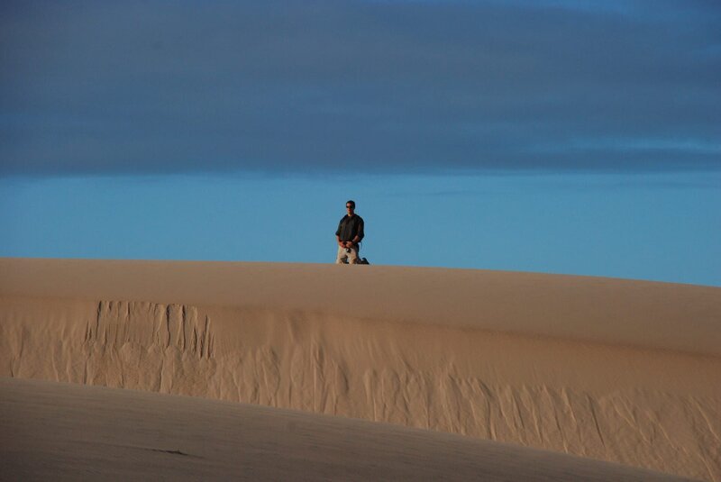 Bear Grylls kneeling on a sand dune, small in the frame. – Bild: Copyright: Discovery Communications, Inc. For Show Promotion Only