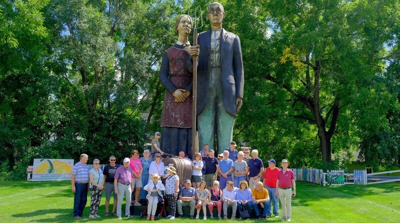 Die Reisegruppe in Anamosa Iowa vor einer Skulptur zur Erinnerung an den Künstler Grant Wood. – Bild: ZDF und NDR/​doc.station/​Volkert Schult.