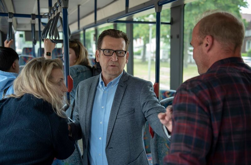 Lars Englen (Ingo Naujoks, M.) hat Mühe, im fahrenden Bus für Ruhe und Ordnung zu sorgen (mit Kompars:innen). – Bild: ARD/​Thorsten Jander