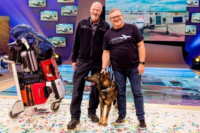Zollbeamter Thomas Dyrda (l.) und sein Spürhund Idris zeigen Elton (r.) bei „1, 2 oder 3“, wie sie am Flughafen Schmuggelware im Gepäck finden. – Bild: ZDF/​Nadine Rupp