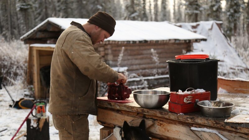 Tyler preparing meat on the table outside his wood cabin. – Bild: Animal Planet /​ Discovery Communications