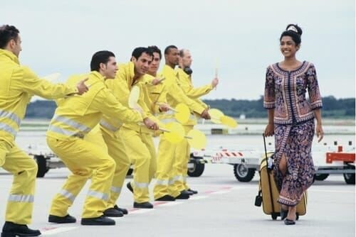 Nisha (Masumi Makhija) als singende Stewardess auf dem Rollfeld mit tanzendem Flughafenpersonal. – Bild: port.hu