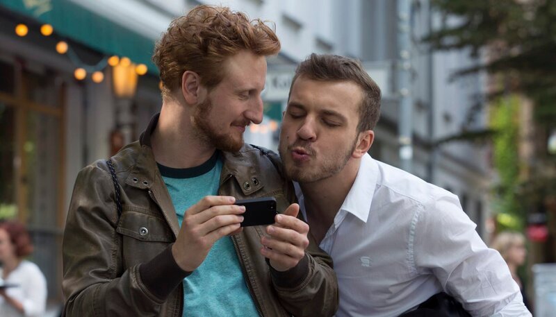 Spaßvogel Mehmet (Edin Hasanovic, rechts) zieht Simon (Michael Kranz) auf, der mit seiner französischen Freundin am Smartphone spricht. – Bild: ARD Degeto/Frédéric Batier Spaßvogel Mehmet (Edin Hasanovic, rechts) zieht Simon (Michael Kranz) auf, der mit seiner französischen Freundin am Smartphone spricht. – Bild: ARD Degeto/Frédéric Batier