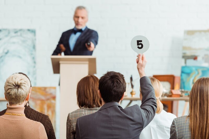 back view of buyer showing auction paddle with number five to auctioneer during auction – Bild: Shutterstock /​ Shutterstock /​ Copyright (c) 2020 LightField Studios/​Shutterstock. No use without permission.