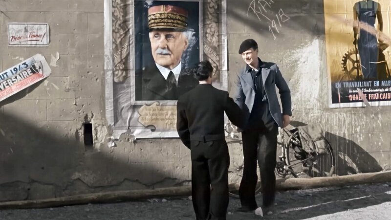 Zwei französische Widerstandskämpfer begrüßen sich vor einem Plakat von Marschall Pétain. – Bild: ORF III