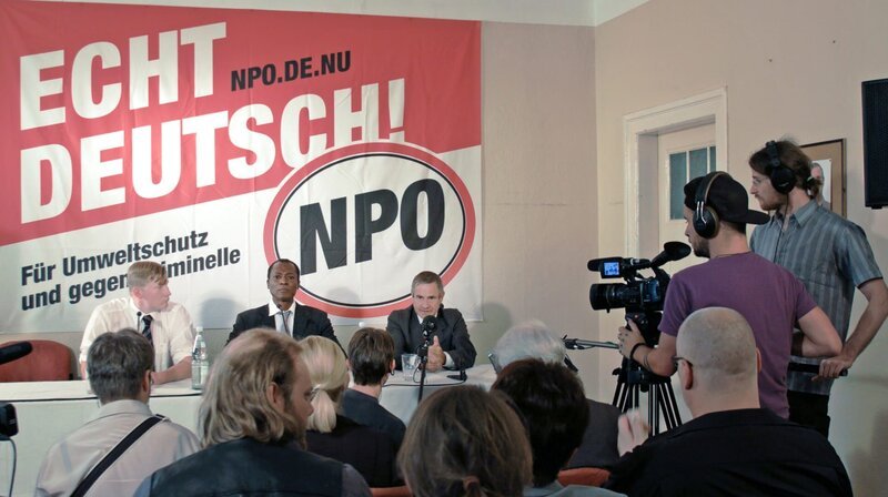 Pressekonferenz: Sikumoya (Aloysius Itoka, mi.) tritt auf einer Pressekonferenz der rechtsextremen Partei NPO auf (mit Bernd-Michael Baier, re. und Christian Weber, li.). – Bild: MDR/Cinemabstruso /Studio Hamburg Enterprises GmbH Pressekonferenz: Sikumoya (Aloysius Itoka, mi.) tritt auf einer Pressekonferenz der rechtsextremen Partei NPO auf (mit Bernd-Michael Baier, re. und Christian Weber, li.). – Bild: MDR/Cinemabstruso /Studio Hamburg Enterprises GmbH