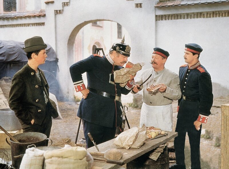 Feldwebel Falkenberg (Rudolf Rhomberg, 2. v. li.) kontrolliert die Erbsensuppe mit Speck, die der Kompaniekoch (Tschik Cajkovski) zubereitet. Lausbub Ludwig (Hansi Kraus, li.) will dem ungehobelten Spieß eine Lektion erteilen. – Bild: port.hu