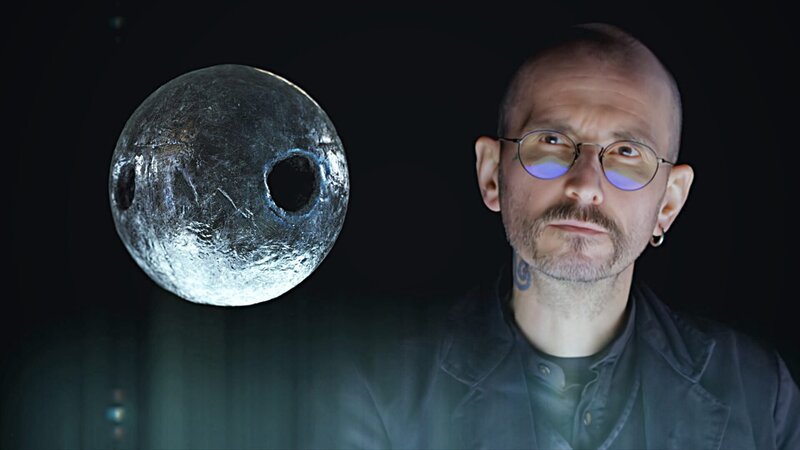 Der Kriminalbiologe Mark Benecke untersucht eine stecknadelkopfgroße, durchlöcherte Metallkugel, die im Körper eines toten Mannes gefunden wurde. – Bild: N24 Doku