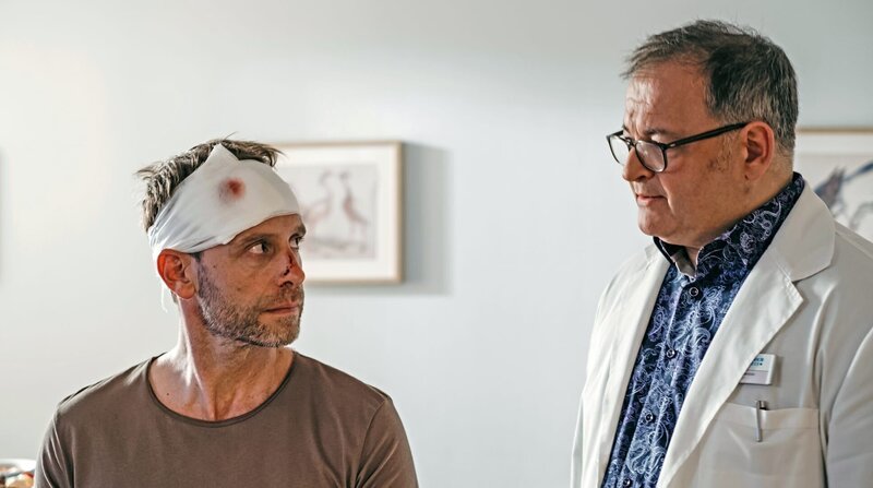 Dr. Kai Hoffmann (Julian Weigend, links) hat sich bei seinem Treppensturz ein Schädel-Hirn-Trauma zugezogen und Ruhe-Pflicht verordnet bekommen. Da Hans-Peter Brenner (Michael Trischan) indirekt Schuld an diesem Unfall ist, soll er nun die Koordination der Notaufnahme übernehmen. Diese ist mehr als überfüllt, da nach einer Massenpanik auf einem Rock-Festival Hunderte von Patienten eingeliefert wurden und werden. – Bild: BR/​MDR/​Saxonia Media/​Robert Strehler