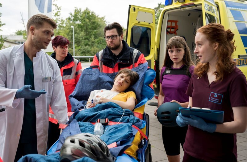 Nele Bader (Sarah Schmidt, M.) wird in Begleitung ihrer Freundin Bianka Schneider (Annemarie Hörold, 2.v.r.) in das Johannes-Thal-Klinikum eingeliefert. Mikko Rantala (Luan Gummich, l.) und Tamar Hummel (Linda Kummer, r.) nehmen die Patientin vom Rettungsdienst (Komparsen, h.) entgegen. – Bild: ARD/​Jens-Ulrich Koch