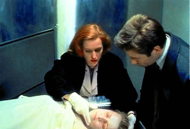 Scully (Gillian Anderson, l.) will Mulder (David Duchovny, r.) eine Phosphoressenz um Mund und Nase einer ermordeten Prostituierten zeigen, die sie bei ihrer ersten Untersuchung entdeckt hat. – Bild: ProSieben MAXX
