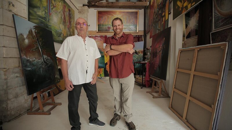 Josh Gates und Michel Becker stehen inmitten der Originalillustrationen für das Golden Owl-Puzzle in Rochefort, Frankreich. – Bild: Discovery Communications