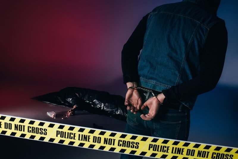 Criminal in handcuffs by body in trash bag behind police tape. – Bild: EdZbarzhyvetsky /​ depositphotos