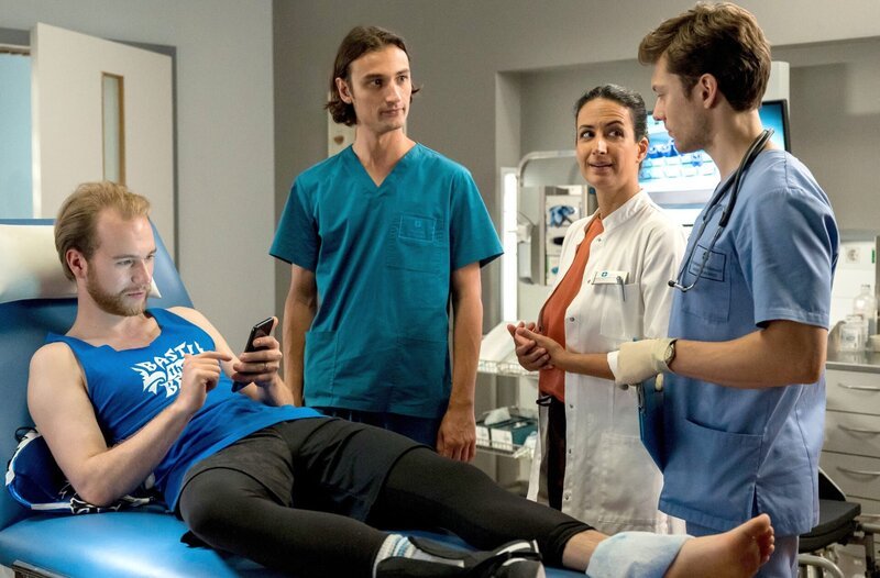 Während der Patient Bastian Kempe mit seinem Handy beschäftigt ist, besprechen Dr. Leyla Sherbaz, Florian Osterwald und Ivo Maric den weiteren Behandlungsverlauf. – Bild: ARD/​Jens-Ulrich Koch