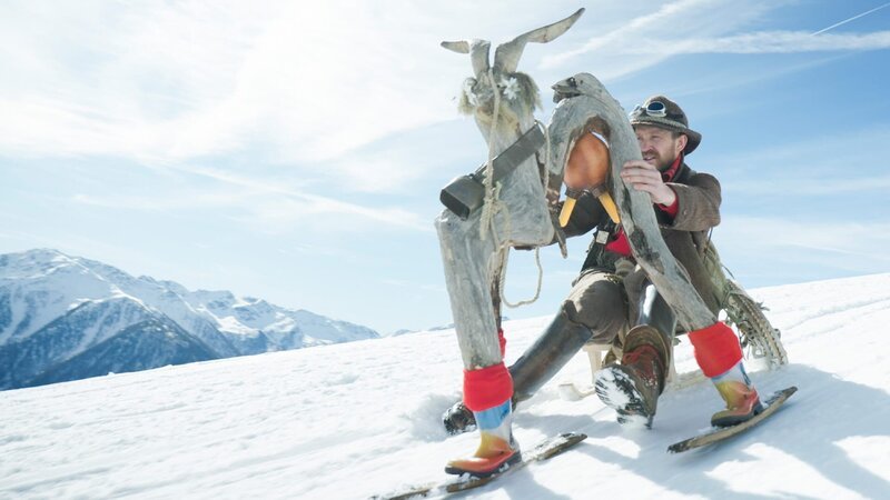 Ein Mann steigt mit einer seltsamen Statue einen schneebedeckten Berg hinab. – Bild: Bergblick