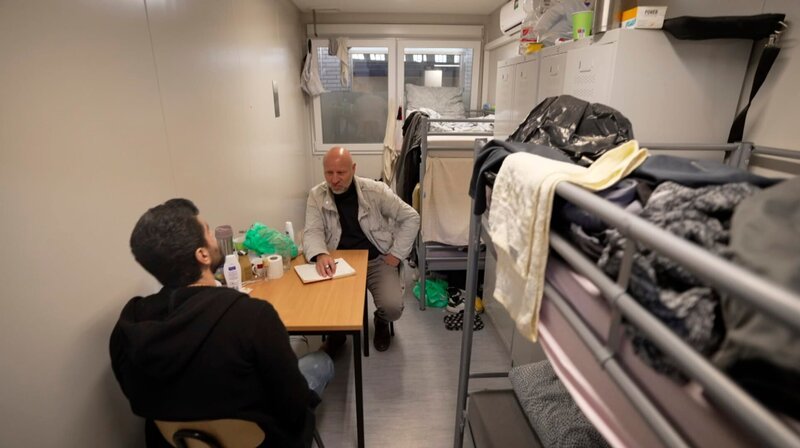 Über irreguläre Migration wird viel gestritten. Der Film begleitet Menschen, die erleben, wie sich ihr Ort verändert – Zugezogene wie Alteingesessene. Und er zeigt Grenzen der Belastbarkeit: in den Oder-Gemeinden ebenso wie in Berliner Massenunterkünften. Reporter Olaf Sundermeyer trifft auch auf Populisten, die mehr Abschiebungen fordern, während sie gleichzeitig Abschiebezentren bekämpfen. – Leben im Container: Aladin Kneher aus Syrien (mit rbb-Reporter Olaf Sundermeyer, re.) lebt mit drei anderen arabischen Flüchtlingen auf 12 qm in der Flüchtlingsunterkunft im Hangar auf dem ehemaligen Flughafen Berlin-Tempelhof, wartet hier auf eine Arbeitsgenehmigung, seit einem Jahr. – Bild: rbb