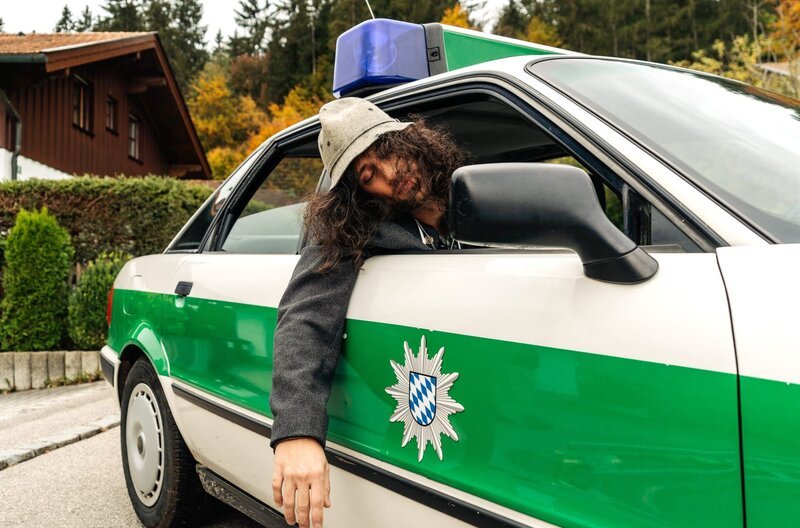 Der Oktoberfest-Tourist Spyros Karamanlis (Nikos Laskaris) ist stark betrunken und wird im Polizeiauto zu seiner Unterkunft gebracht. – Bild: ARD/​Max Bublak/​Max Bublak