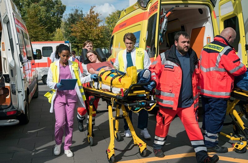 Nach einem Massenunfall werden mehrere Patienten am Johannes-Thal-Klinikum eingeliefert. Emma Jahn (Elias Agbaglah, l.) und Mikko Rantala (Luan Gummich, 3.v.r. mit Komparsen) kümmern sich um Frieda Kantak (Pauline Rénevier, M.). – Bild: Jens Ulrich Koch