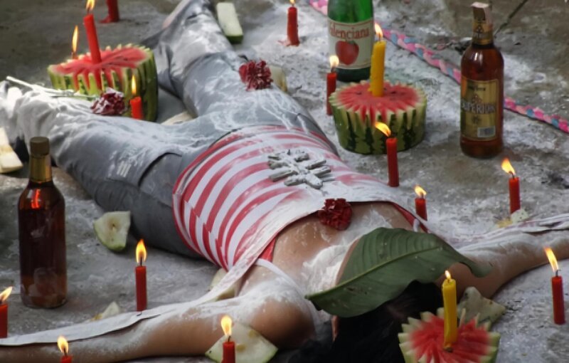 Woman lies flat on the ground surrounded by candles, fruit and alcohol with amulet on her chest and a leaf across her face and covered in white powder. – Bild: National Geographic Society