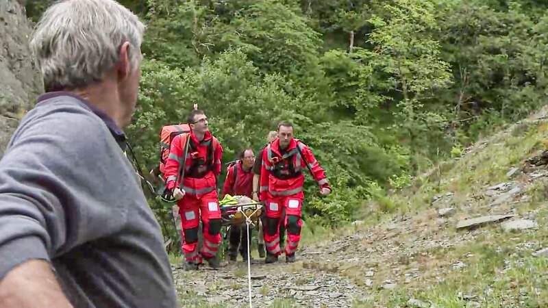 Tyrone, Paul und MRT tragen einen Patienten einen felsigen Hügel hinauf. – Bild: Discovery Communications