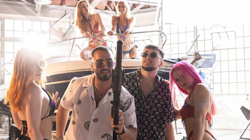 Die Rapper (Simon Nouri, li. und Cemal Cakiroglu, re.) posen mit Waffen und leicht bekleideten Frauen für ihr Musikvideo. – Bild: ARD Degeto/​Majestic Film/​Dor Film/​Viafilm/​Scarlett Werth