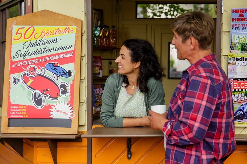 Fritz Fuchs (Guido Hammesfahr) verrät Yasemin (Sanam Afrashteh), dass er größte Lust hätte, selbst noch einmal an einem Seifenkistenrennen teilzunehmen. – Bild: Andrea Hansen