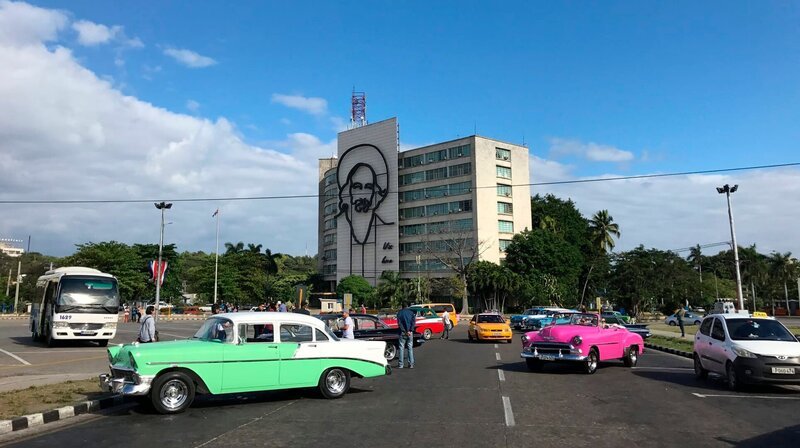 Bunte Oldtimer parken vor dem Informationsministerium mit einem Porträt von Camilo Cienfuegos in Havanna auf Kuba. – Bild: BR/​Gerrit Mannes