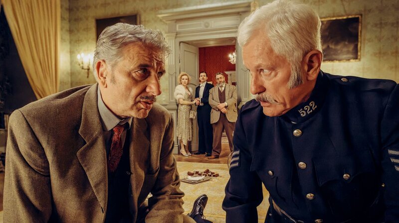 Chief Inspector Francis Lightmyer (Udo Wachtveitl, links vorne) und Constable Ivor Partridge (Miroslav Nemec, rechts vorne) untersuchen Arthur im Kaminzimmer. Lady Bantam (Sunnyi Melles), Charles (Ferdinand Hofer) und Dr. Mallard (Alexander Hörbe, rechts hinten) beobachten sie. – Bild: HR/​BR/​Bavaria Fiction GmbH/​Hendrik Heiden